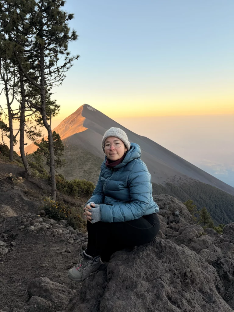 Audrey en doudoune et bonnet, assise sur un rocher au sommet d'une montagne avec un volcan en arrière-plan au coucher du soleil